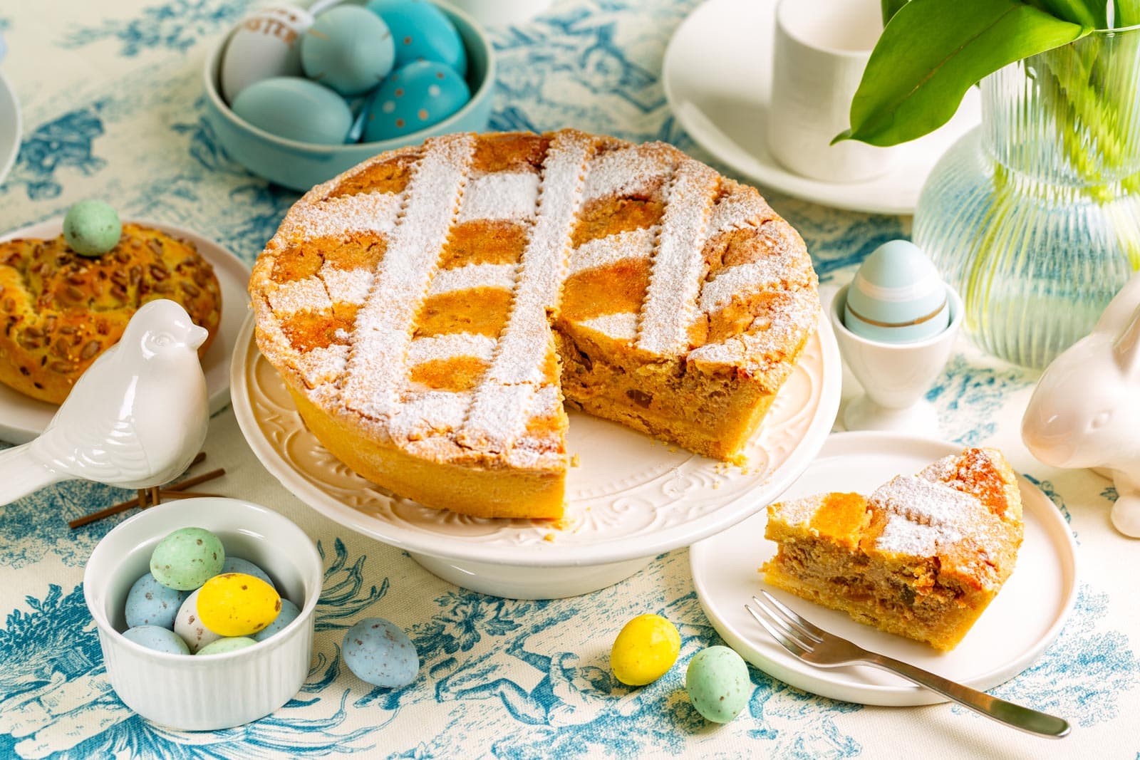 shutterstock 2580640835 ©olgabombologna italian easter cake pastiera napoletana shortcrust pie with ricotta cheese filling and cooked grains colorful eggs buns vase with tulips